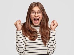 Glad European woman wears spectacles, clenches fists in excitement, wears round spectacles and casual sweater, exclaims with happiness, has dark hair and pleasant appearance, models over white wall