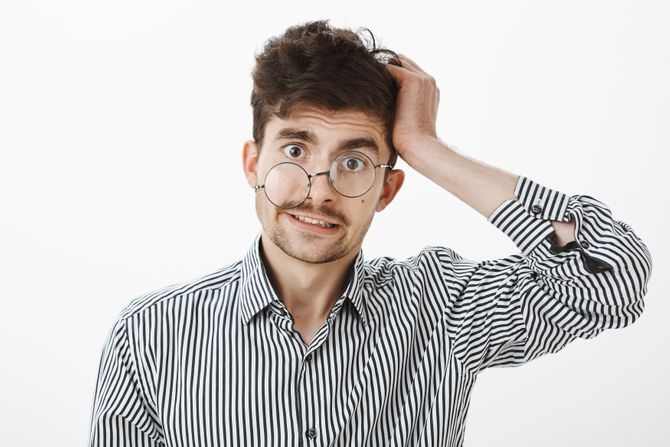 Guy after hangover lost memory. Confused questioned messy male in striped shirt, scratching hair and looking unaware and clueless at camera with glance from under glasses, standing over gray wall